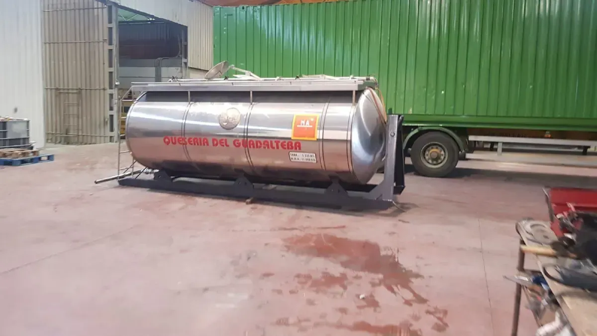 Un remolque cisterna plateado volcado junto a un contenedor de transporte verde en un patio industrial.