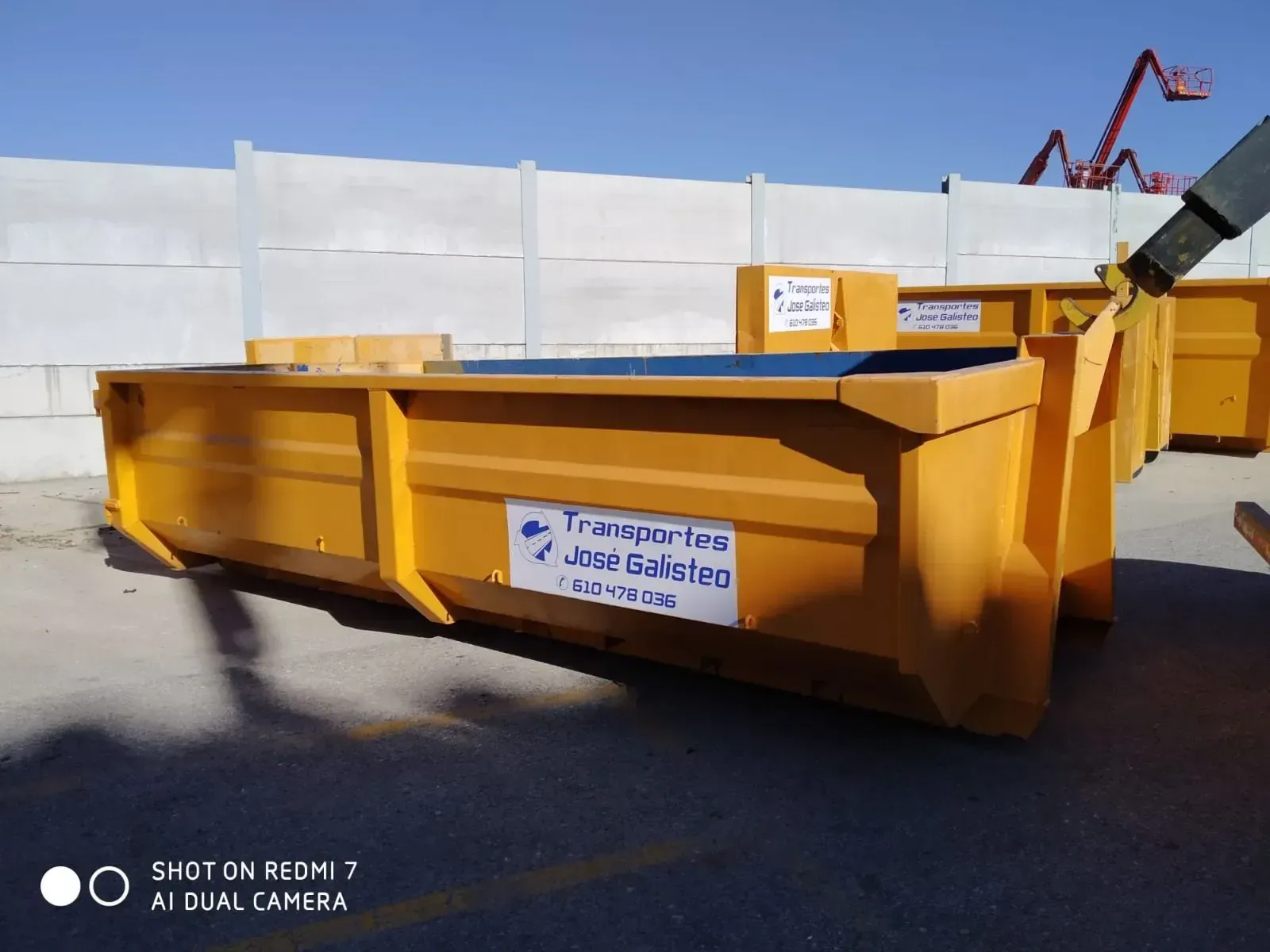 Un contenedor de obra amarillo con una etiqueta, situado al aire libre sobre el pavimento junto a una valla.