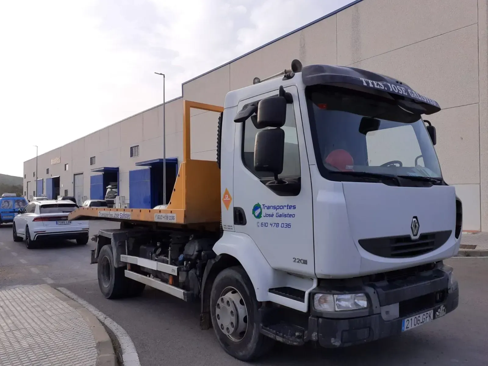 Camioneta blanca estacionada junto a una zona de carga de un almacén, con contenedores azules y coches al fondo.