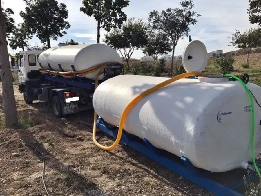 Tanques cilíndricos blancos sobre remolques azules en un campo de tierra, con mangueras conectadas y árboles al fondo.