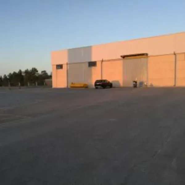 Aparcamiento vacío junto a un almacén de color beige al atardecer, con algunos vehículos estacionados.