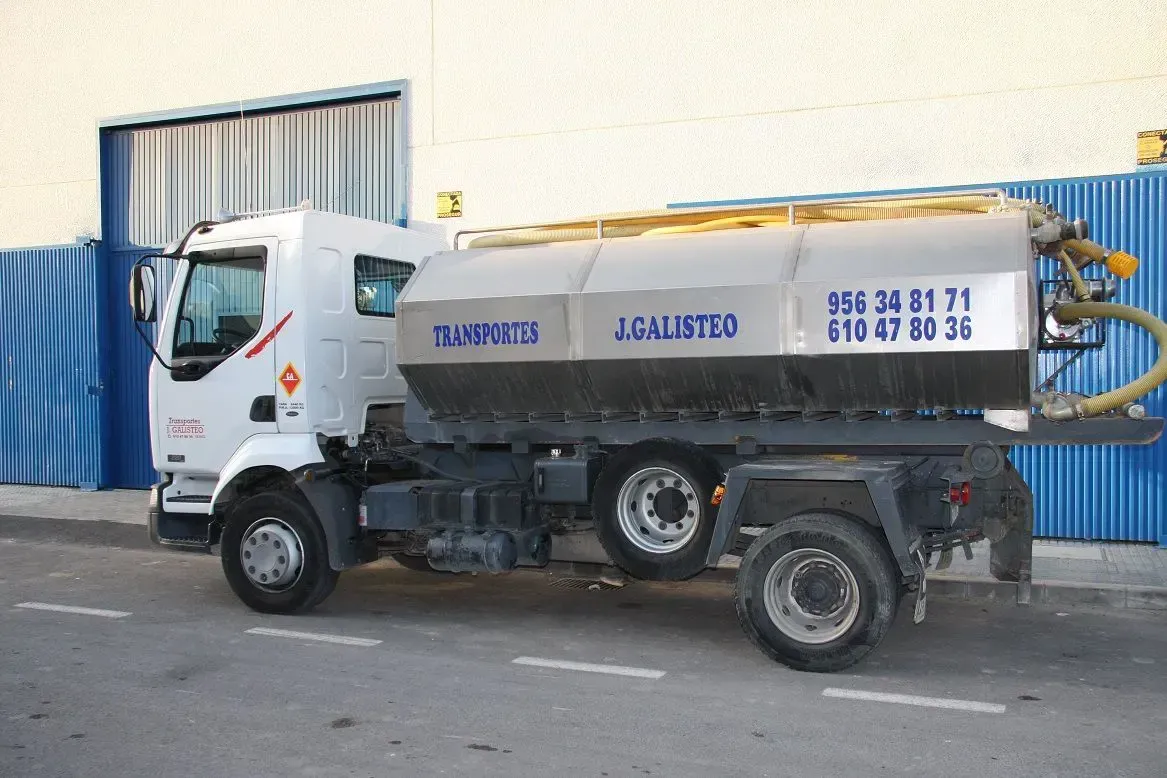 Camión utilitario blanco con una cisterna plateada estacionado junto a contenedores industriales azules.