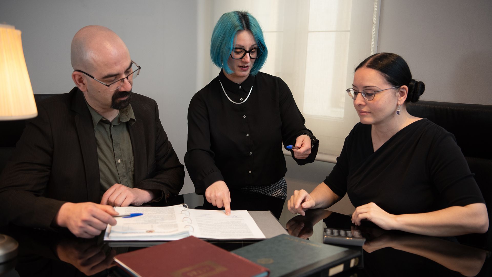 Tres personas revisan documentos en una mesa. Una mujer de pelo azul señala con el dedo, mientras otras dos observan.