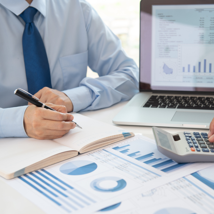 Hombre con camisa azul, usando una calculadora, un cuaderno y una computadora portátil con gráficos financieros.