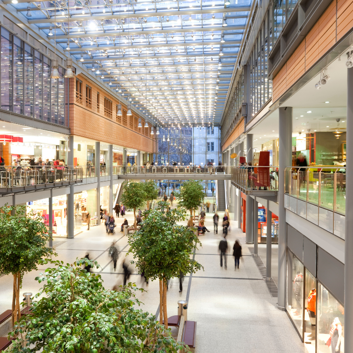 Vista interior de un moderno centro comercial con luz natural, múltiples niveles, tiendas y figuras borrosas caminando.