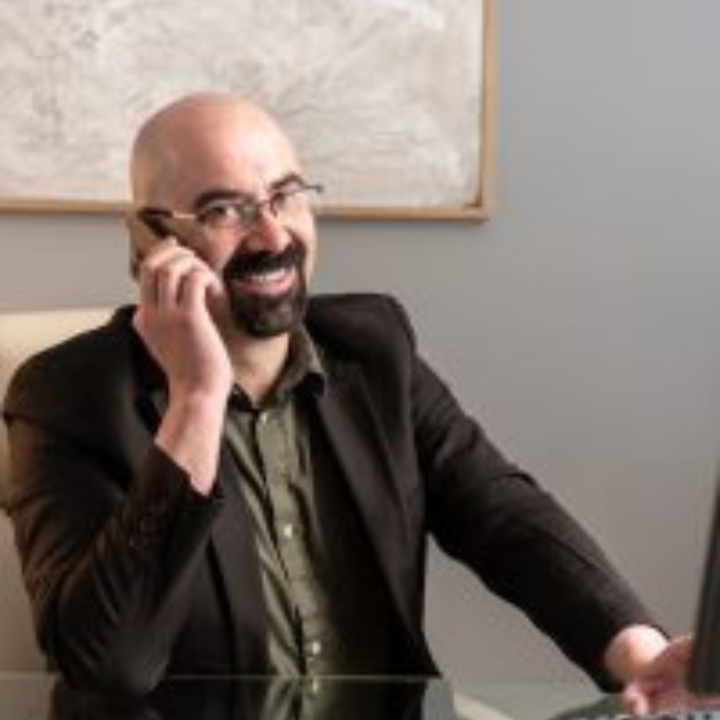 Hombre hablando por teléfono, sonriendo, con gafas y chaqueta, sentado en un escritorio con una computadora portátil.