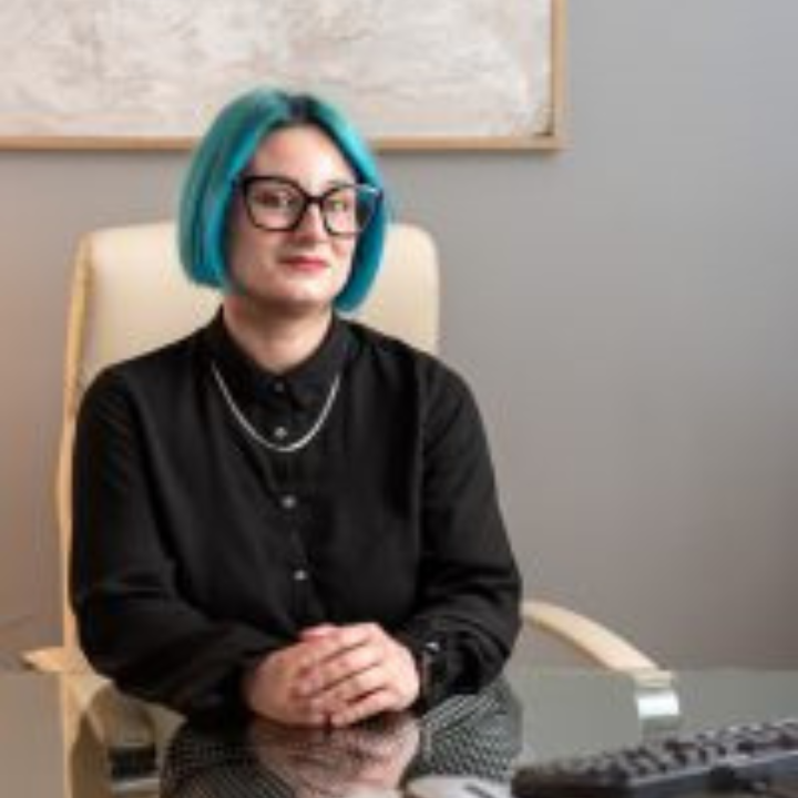 Mujer con cabello azul y gafas sentada en un escritorio, vistiendo una camisa negra, mirando al espectador.