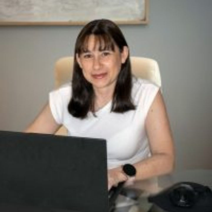 Mujer con cabello oscuro, top blanco, sentada en un escritorio con una computadora portátil y un teléfono, en el interior.