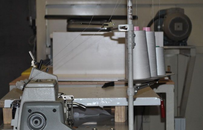Máquina de coser con conos de hilo blanco, banco de trabajo, motor y tela en un entorno de taller.