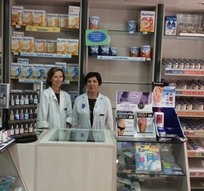 Dos mujeres con batas blancas detrás de un mostrador de farmacia con estantes de productos.