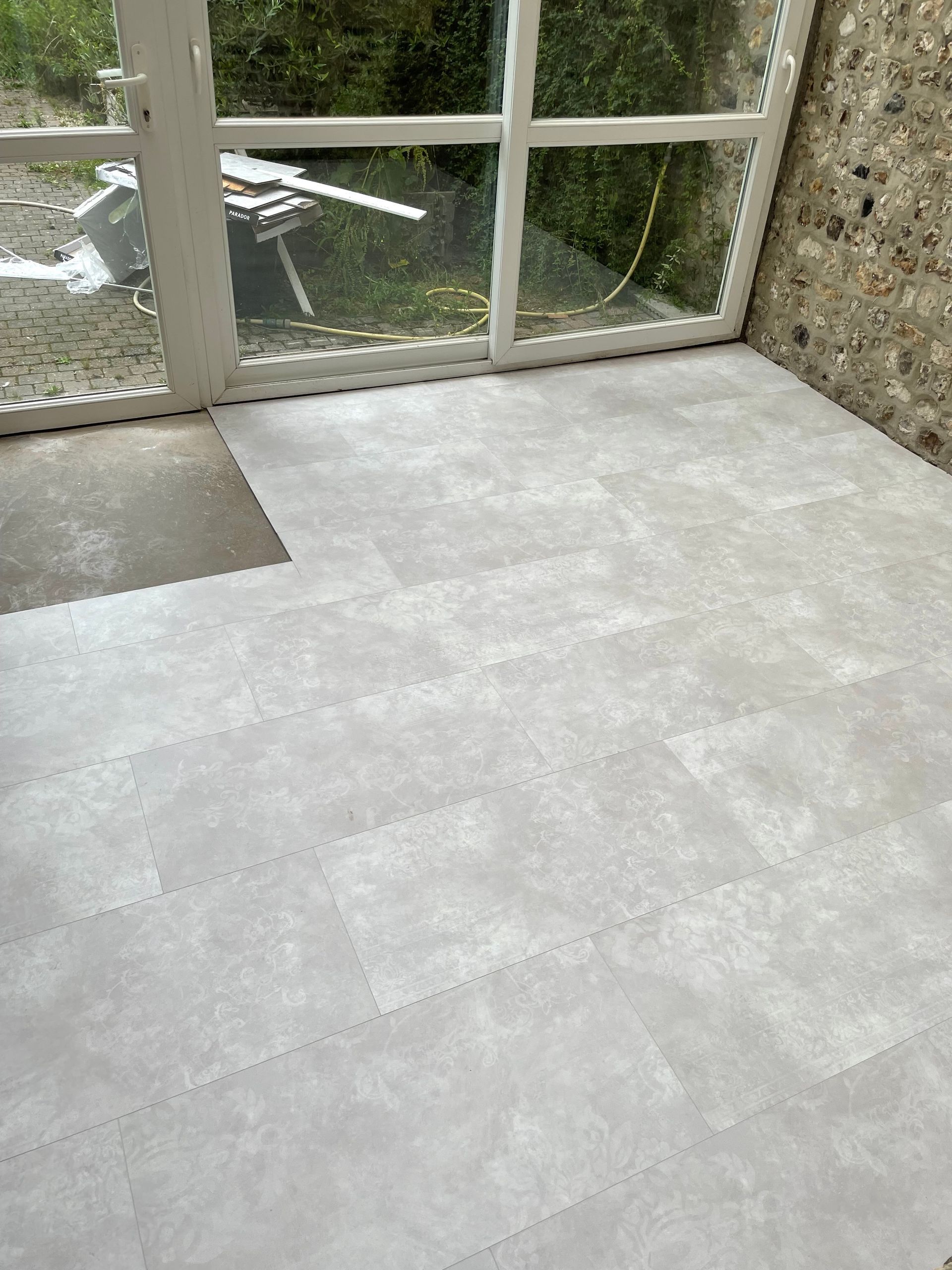 Carrelage de sol gris clair installé dans une véranda avec fenêtre et mur en pierre.
