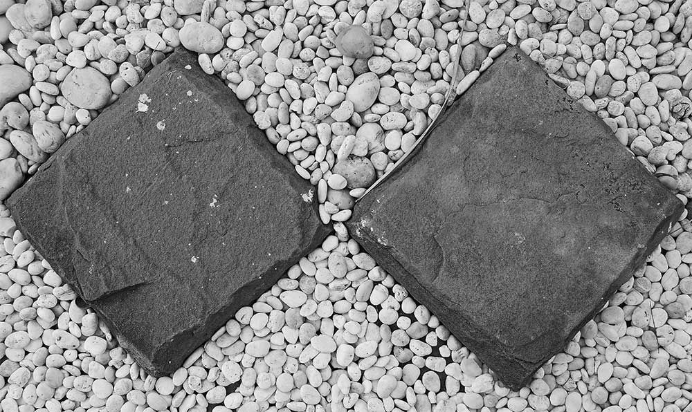 Dos piedras cuadradas y oscuras sobre un lecho de guijarros pequeños y de color claro.