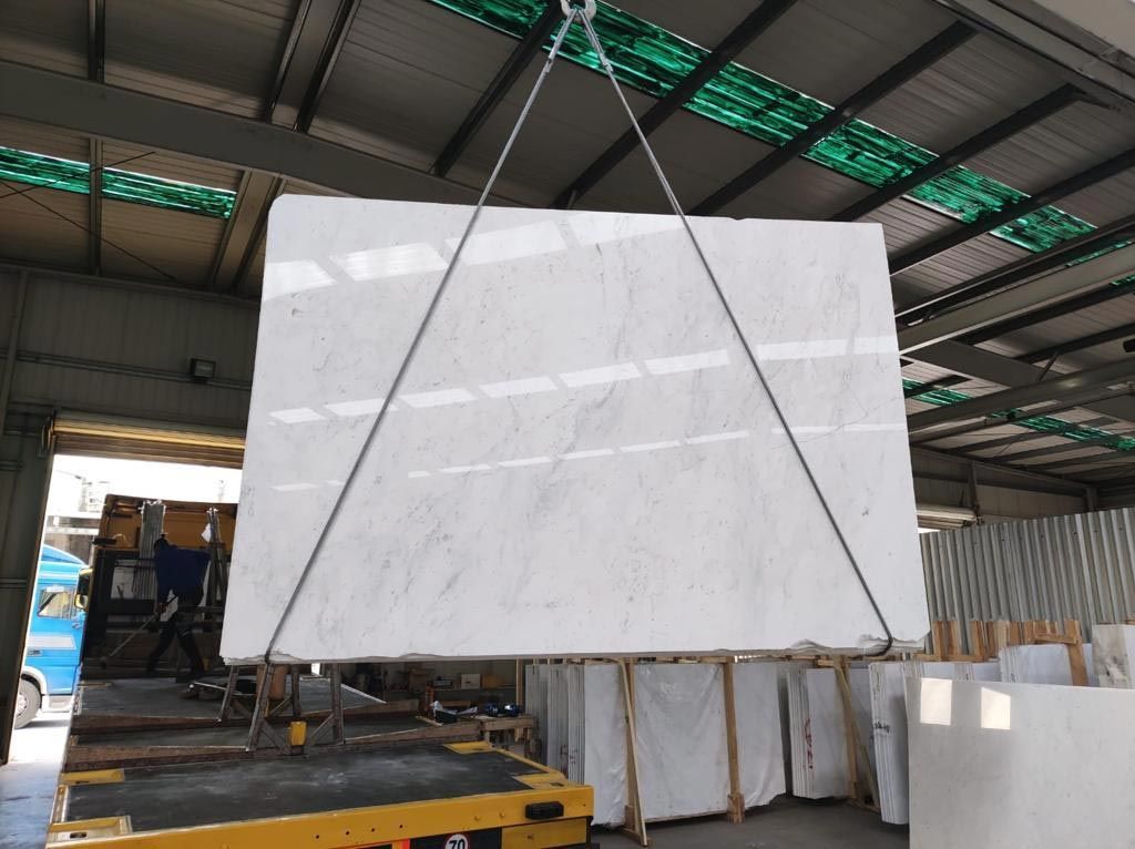 Gran losa de mármol blanco suspendida por cadenas en un almacén, siendo levantada.