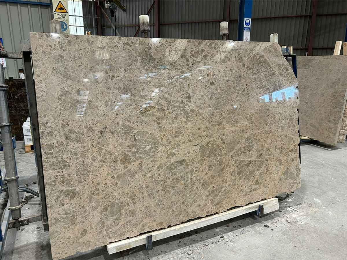 Gran losa de mármol pulido con vetas de color tostado y marrón, apoyada sobre un soporte en un almacén.