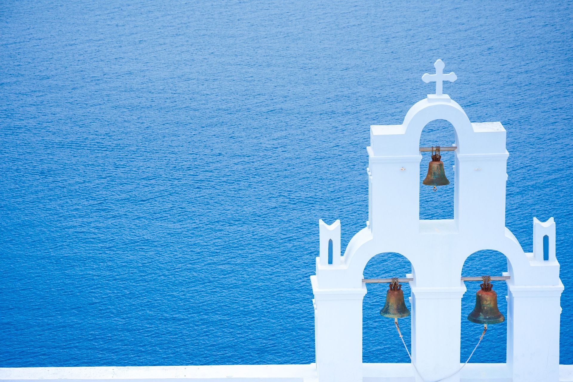 A white church with bells hanging from it overlooking the ocean.