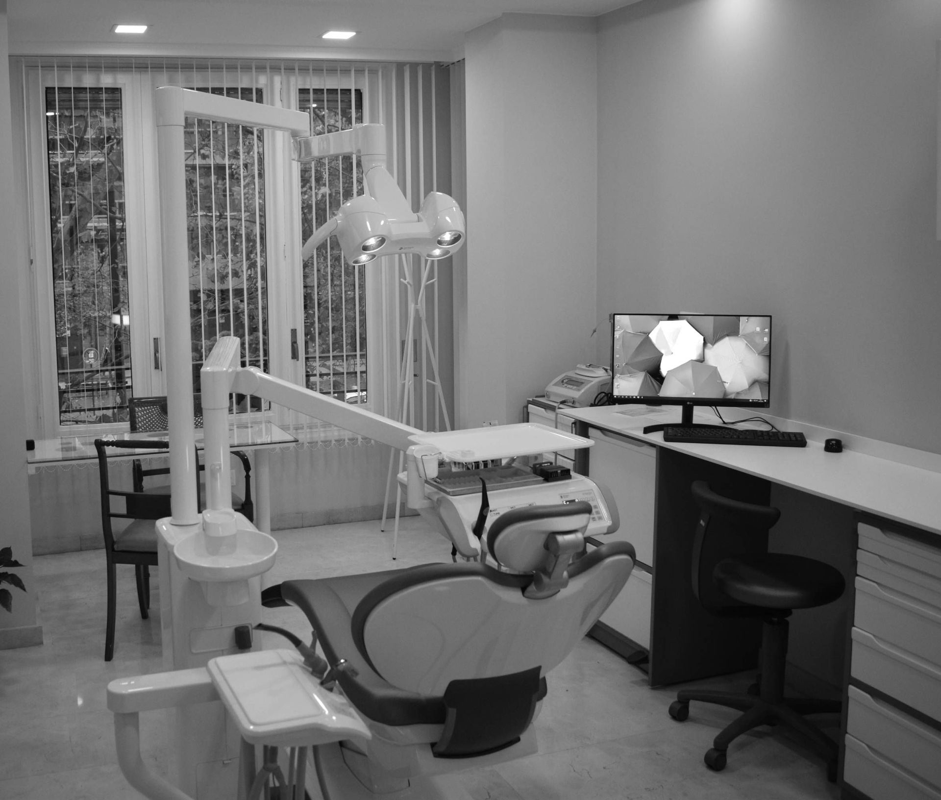Sala de examen dental en blanco y negro; silla, luz, computadora, ventana con persianas.