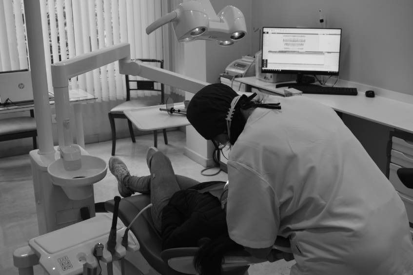 Un dentista con mascarilla examina a un paciente en un sillón. Consultorio, blanco y negro.