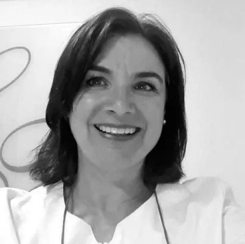 Mujer sonriente con cabello oscuro, vestida con una chaqueta blanca, en el interior.