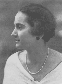 Mujer de perfil, con collar de perlas, cabello corto y ropa de color claro, posiblemente al aire libre.