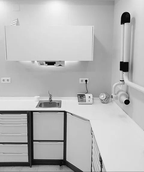 Espacio de trabajo en clínica dental: muebles blancos, lavabo, encimera, equipamiento y brazo de rayos X montado en la pared.