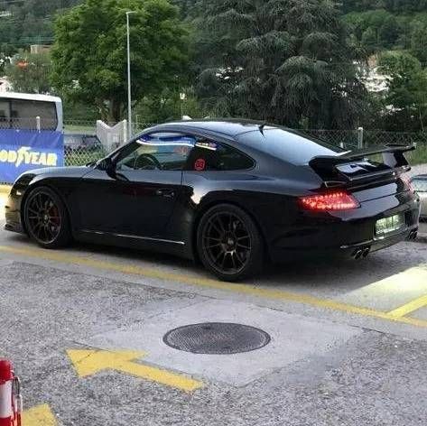 Una Porsche 911 GT3 nera con alettone posteriore parcheggiata su una superficie asfaltata con segnaletica direzionale gialla.