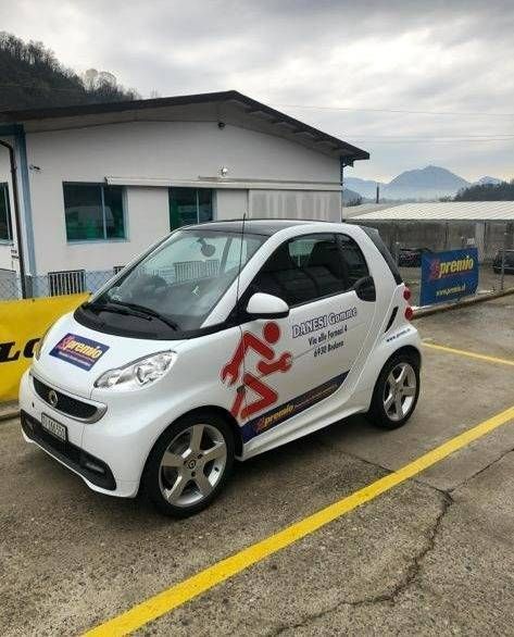 Una Smart bianca parcheggiata fuori da un'officina, con il logo e il marchio di un meccanico di colore rosso sulla fiancata.