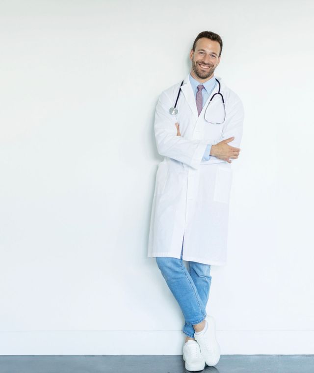 Lächelnder Arzt mit verschränkten Armen, an einer weißen Wand lehnend, mit Laborkittel, Stethoskop, Jeans und Turnschuhen.