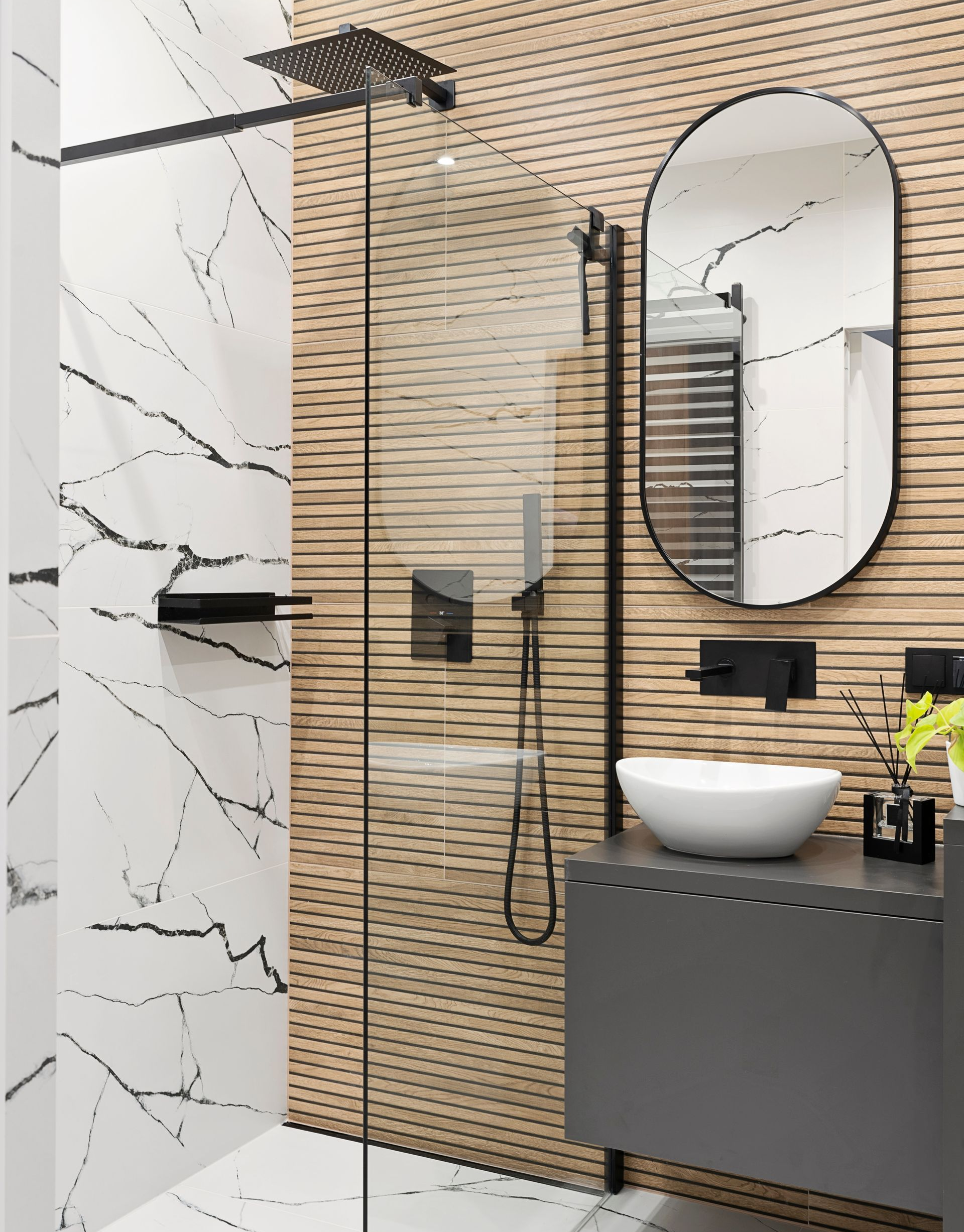 Salle de bains moderne avec des parois en marbre et en bois ; robinetterie noire, miroir ovale, meuble lavabo gris et porte de douche en verre.