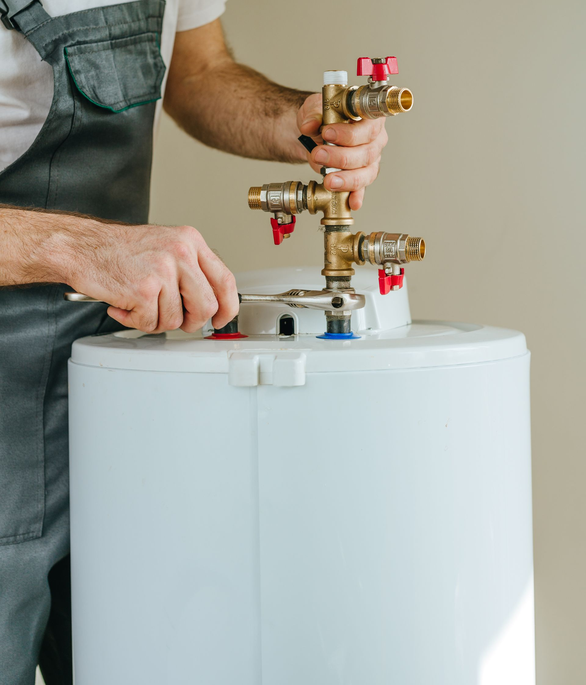 Un plombier en salopette grise utilise une clé sur les vannes d'un chauffe-eau.
