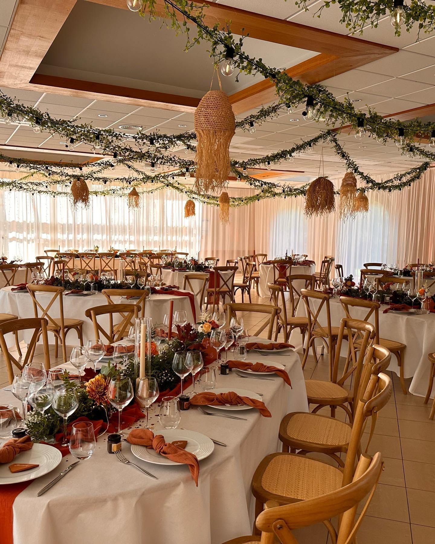 Une salle de réception avec des tables nappées de blanc, des chaises en bois et une décoration végétale suspendue.