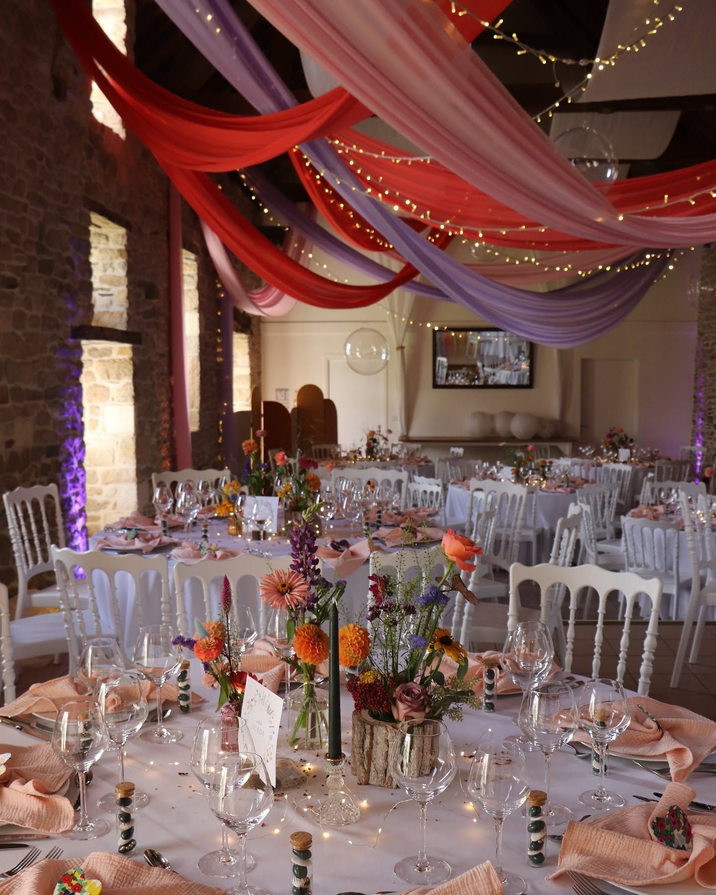 Salle de réception avec des tables rondes, chaises blanches et plafond drapé de rouge, rose et violet.