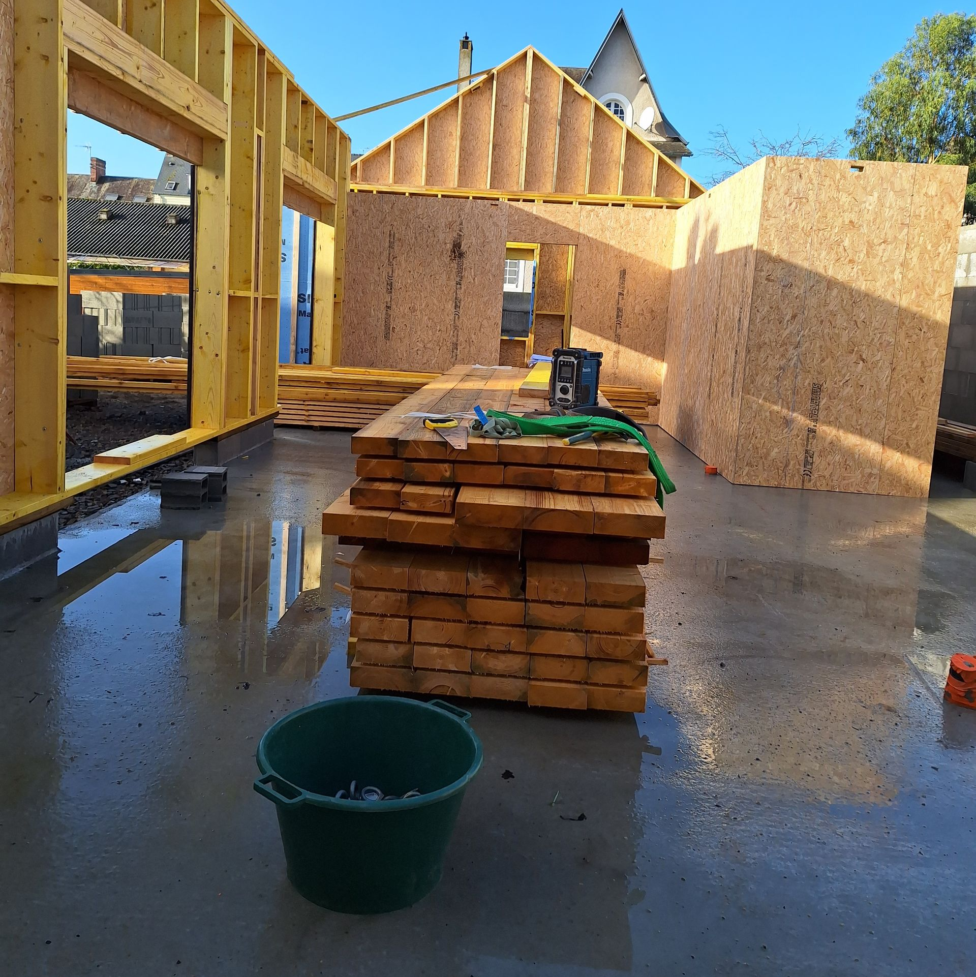 Chantier : murs à ossature en bois en cours de construction sur une dalle de béton, tas de bois au premier plan, seau vert.