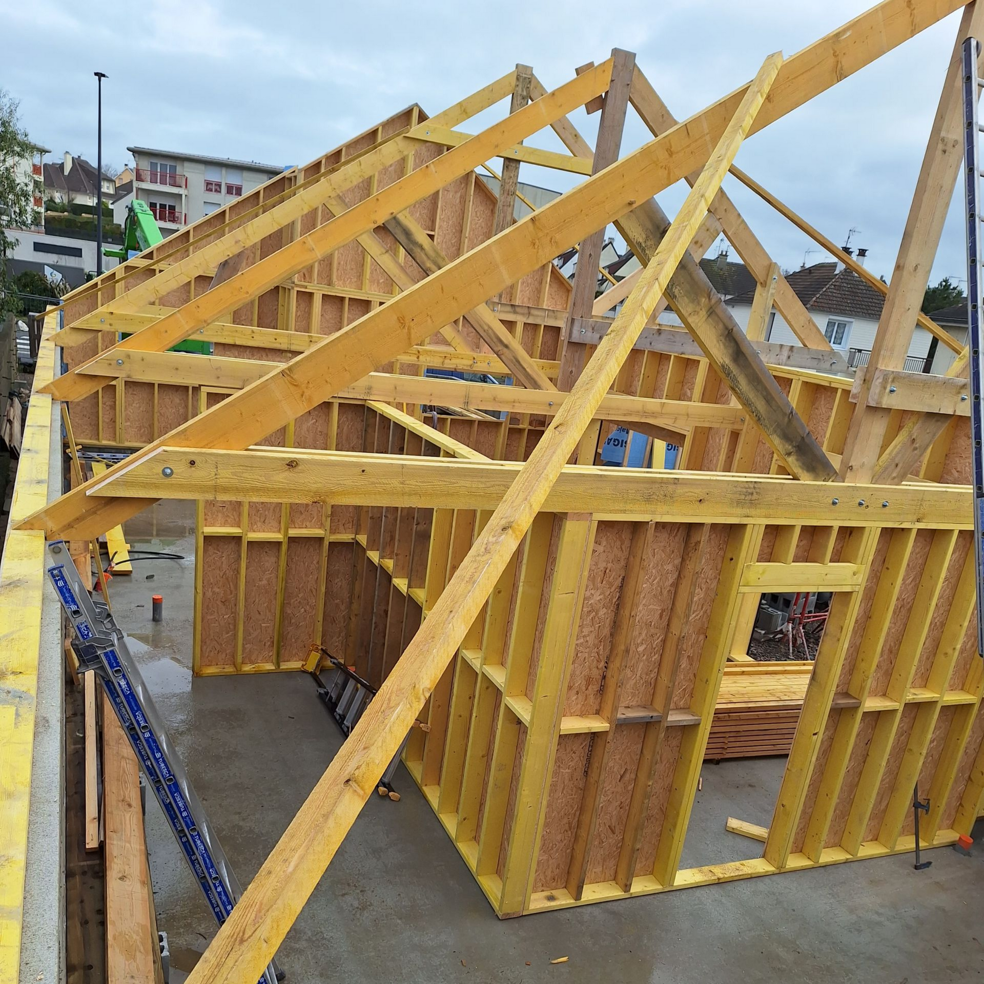 Ossature en bois d'un bâtiment en construction, avec murs à ossature jaune, poutres apparentes et fondation en béton.