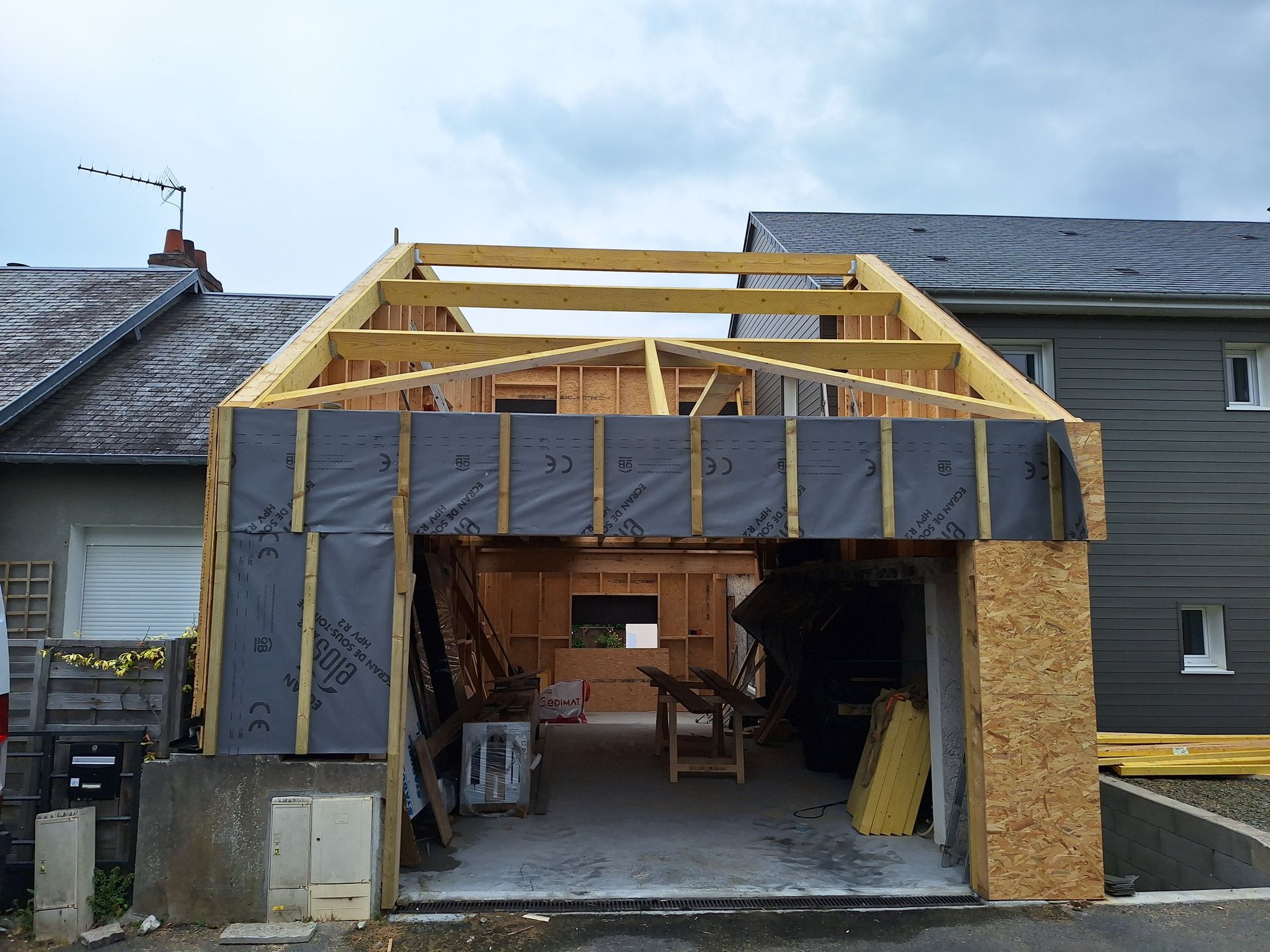 Extension de garage en construction, avec ossature en bois, revêtement noir, intérieur ouvert et ciel nuageux.