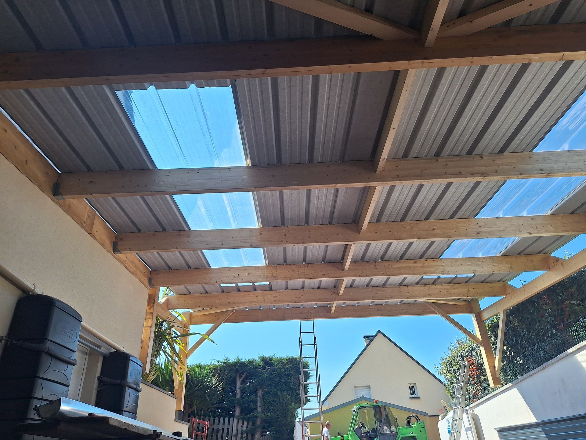 Abri d'auto à ossature en bois avec tôle ondulée et panneaux transparents ; couvrant partiellement un espace extérieur.