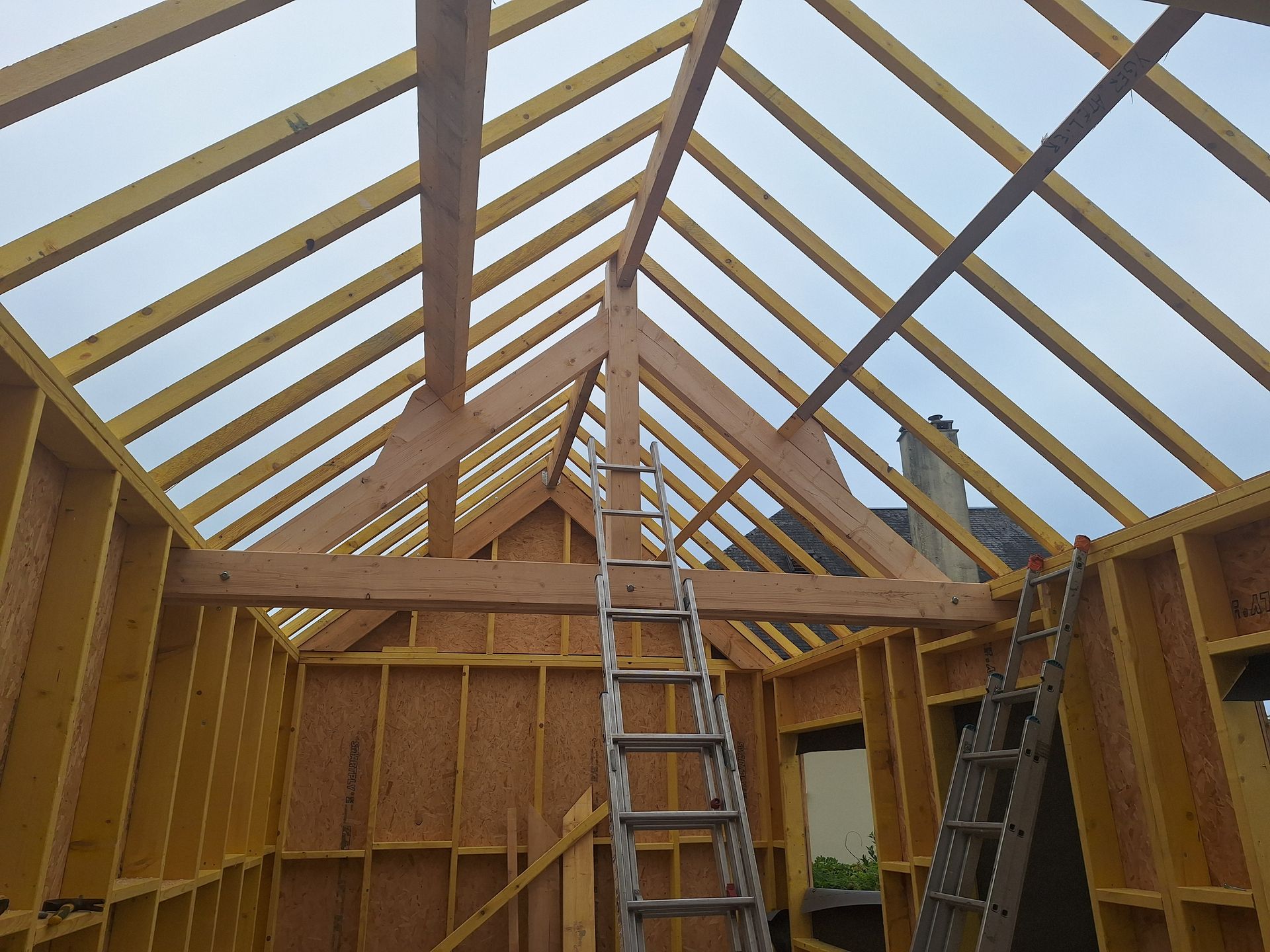 Ossature de maison en bois en construction ; chevrons, poutres, échelle et murs visibles.