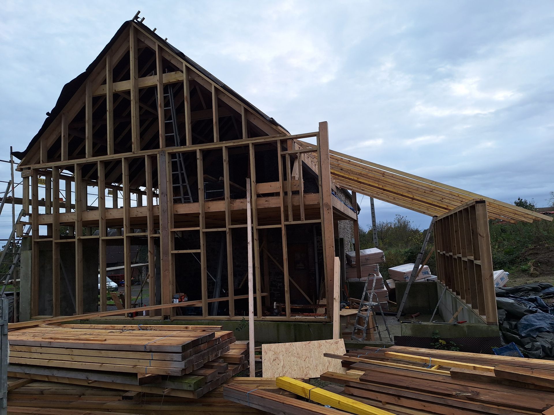 Structure en bois en construction avec charpente apparente et un ciel nuageux en arrière-plan.