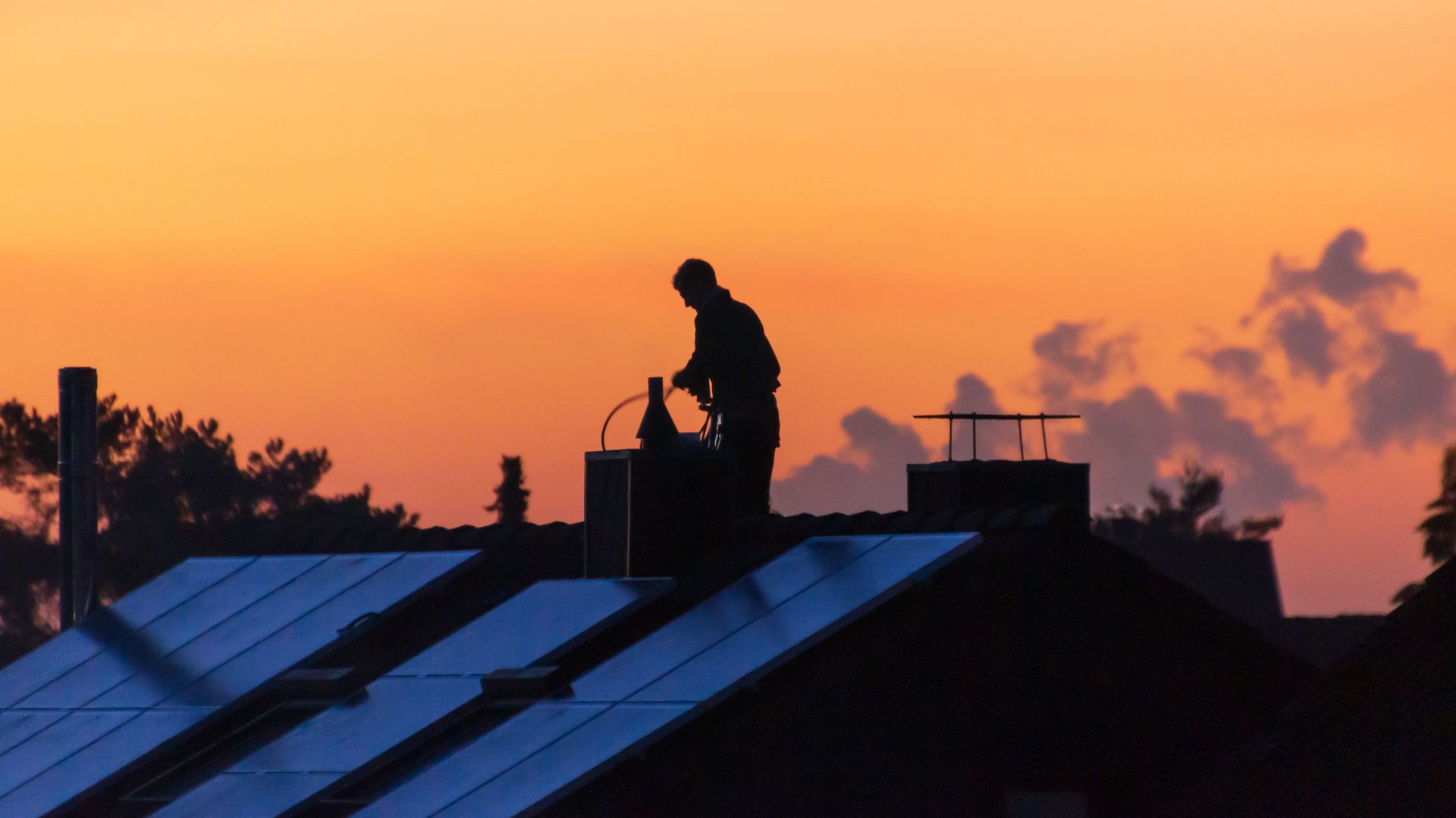 Silhouette d'une personne travaillant sur un toit près d'une cheminée. Panneaux solaires et coucher de soleil orange en arrière-plan.