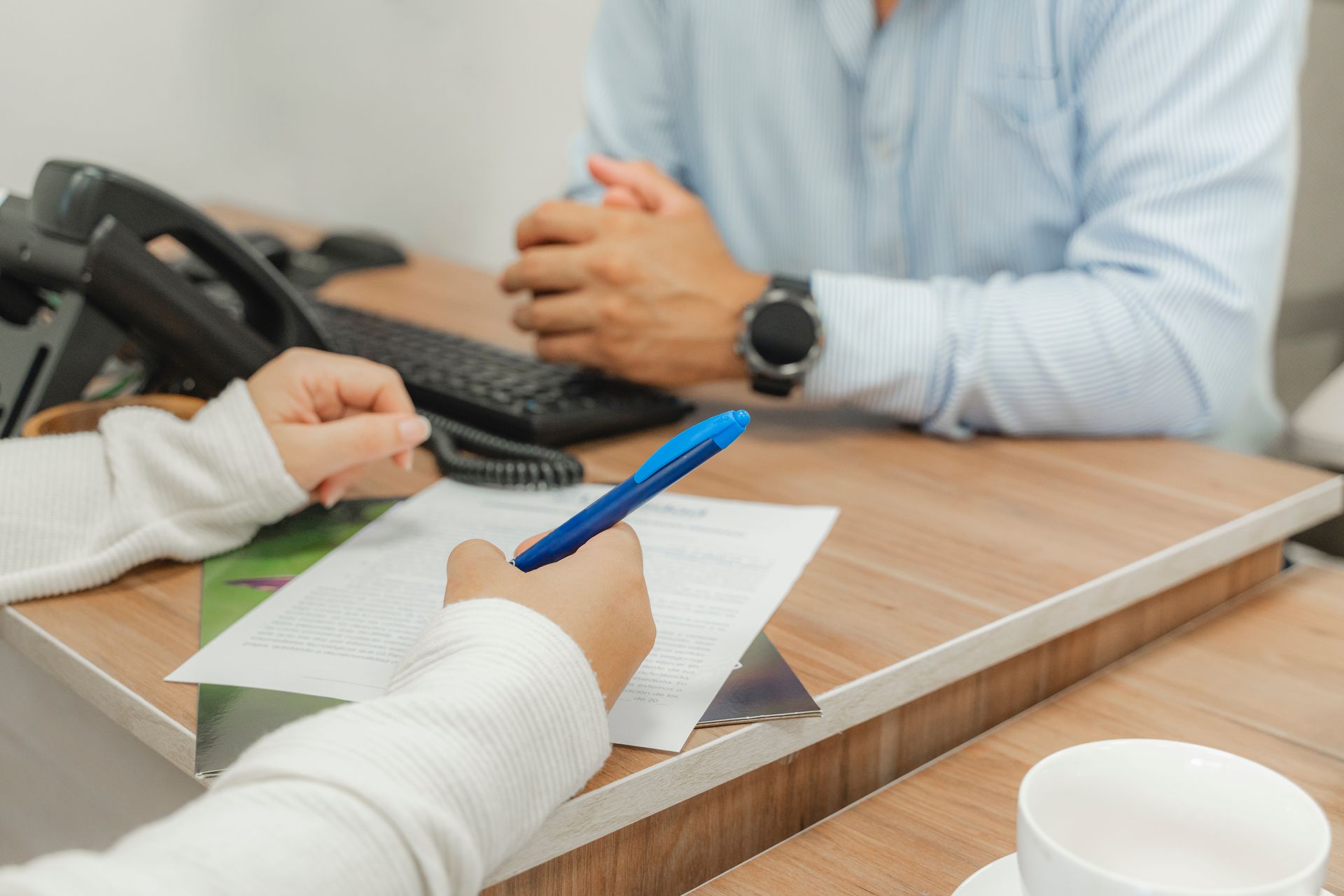 Des mains écrivent sur une feuille de papier à un bureau pendant une réunion avec un homme en chemise bleu clair assis en face d'elles.