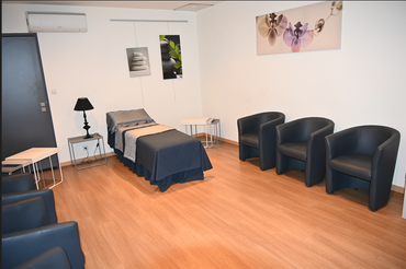 Chambre funéraire vide avec un lit, des fauteuils noirs, des tables d'appoint et des tableaux au mur.