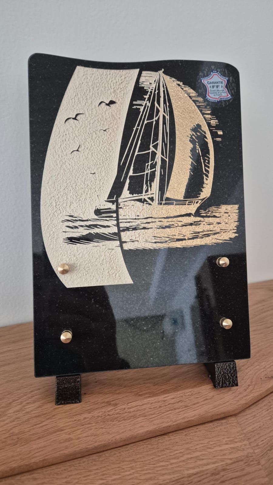 Plaque noire gravée d'un voilier beige, montée sur un petit support sur une surface en bois.