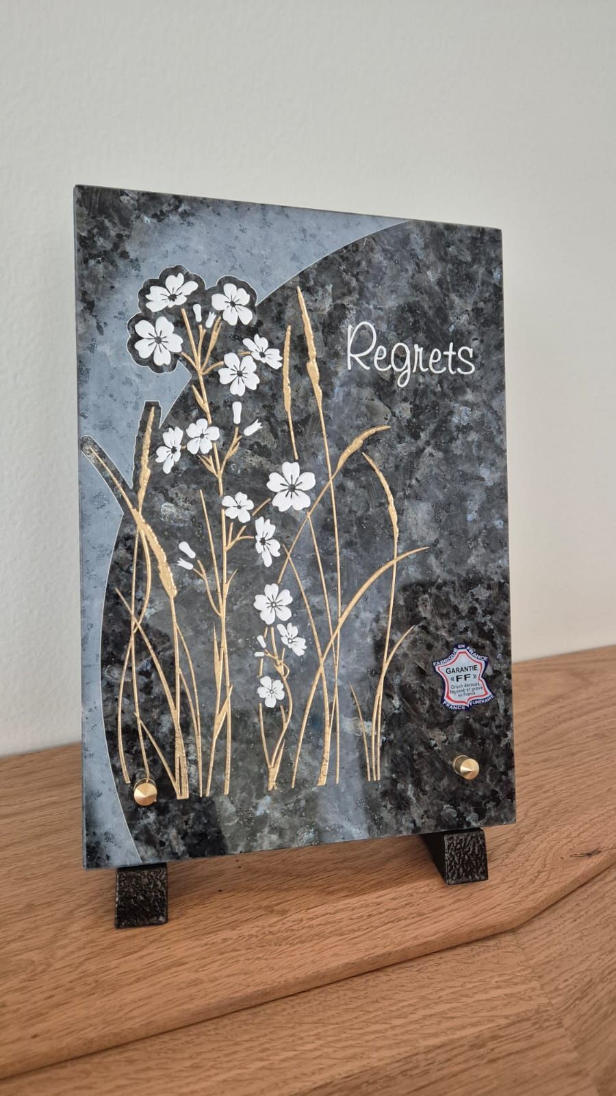 Plaque décorative à motifs floraux sur une surface en bois, avec des fleurs blanches et des tiges dorées sur un fond texturé foncé.