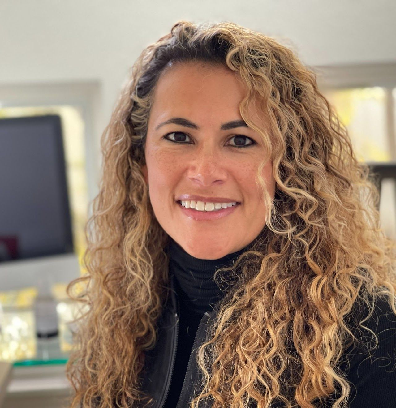 A woman with curly hair is smiling in front of a computer monitor