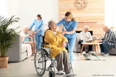 Pflegeheim: Pflegekraft hilft älterer Frau im Rollstuhl; andere Bewohner entspannen sich in der Nähe.