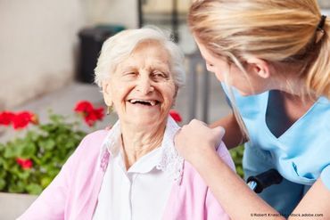 Ältere Frau lächelt, wird im Freien von einer Krankenschwester betreut, rosa Pullover, Blumen im Hintergrund.