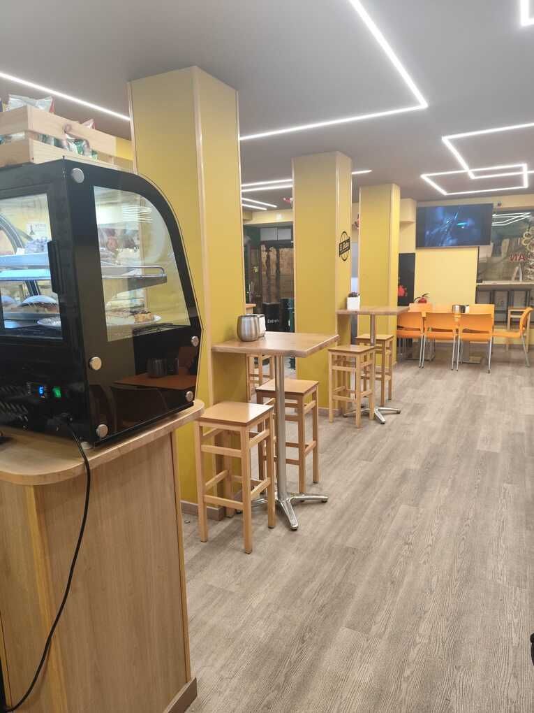 Interior de una cafetería bien iluminada con columnas amarillas, taburetes y mesas de madera. A la izquierda hay una vitrina.