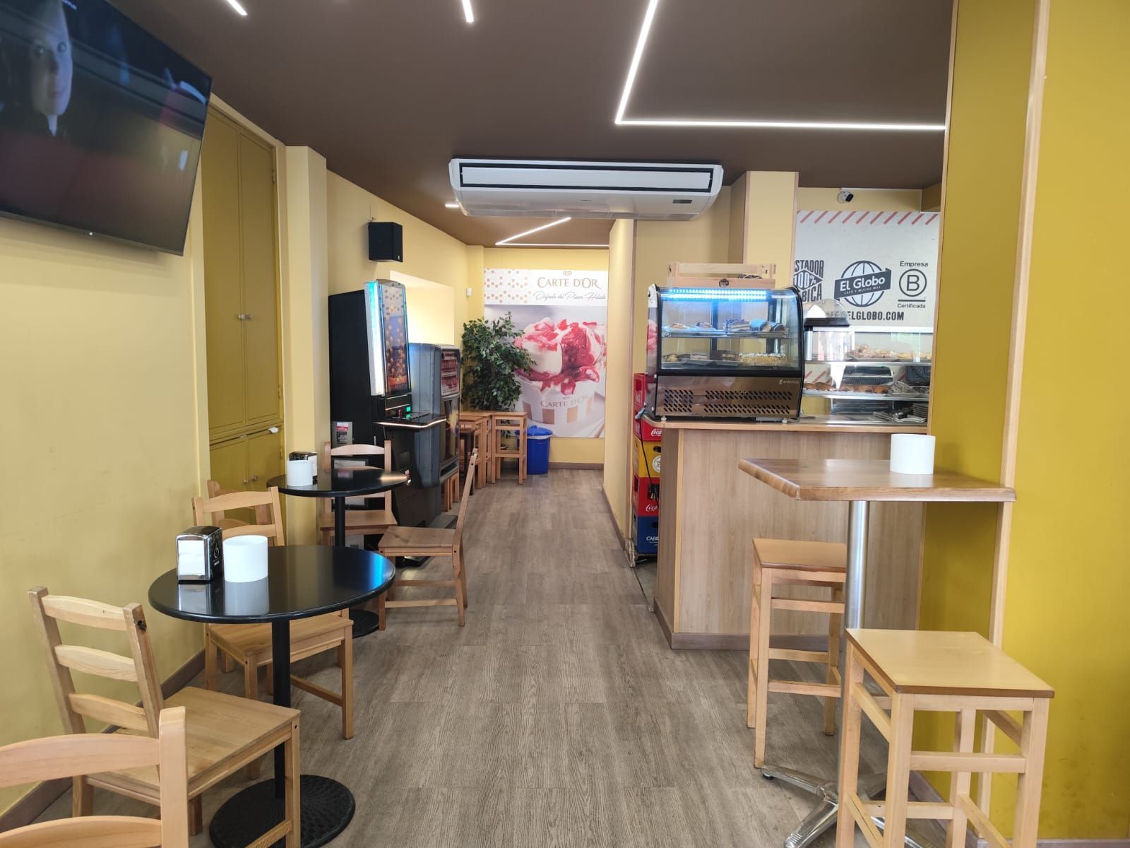 Interior de una cafetería con mesas, sillas, cafetera y vitrina. Paredes amarillas, suelo de madera y un televisor.