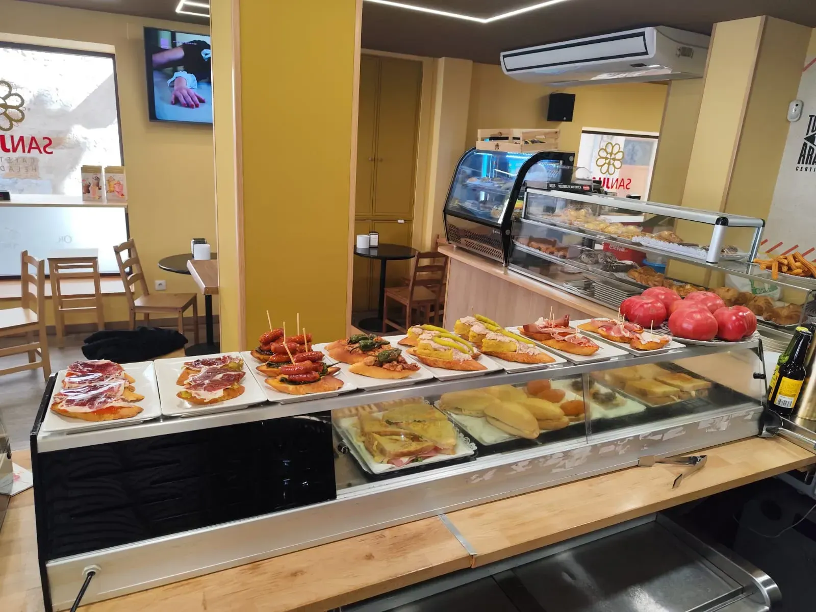 El mostrador de un restaurante ofrece diversas tapas y productos horneados. Paredes amarillas, sillas de madera y una pequeña zona para sentarse.