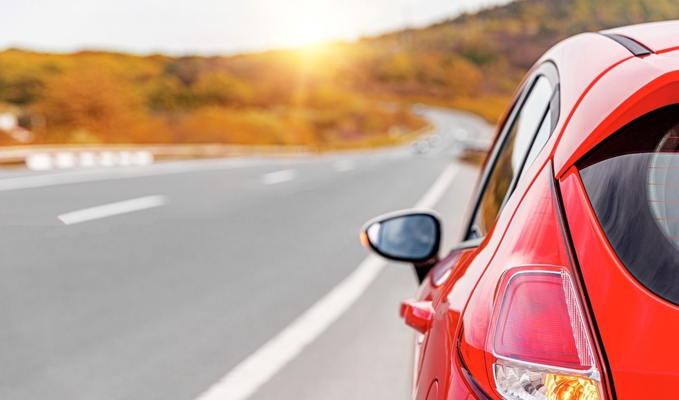 Coche rojo aparcado al costado de una carretera sinuosa con un fondo soleado y borroso de árboles y cielo.