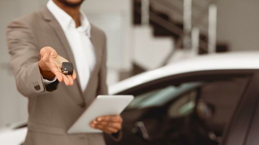Vendedor de coches con traje sosteniendo las llaves del coche y una tableta junto a un coche blanco.